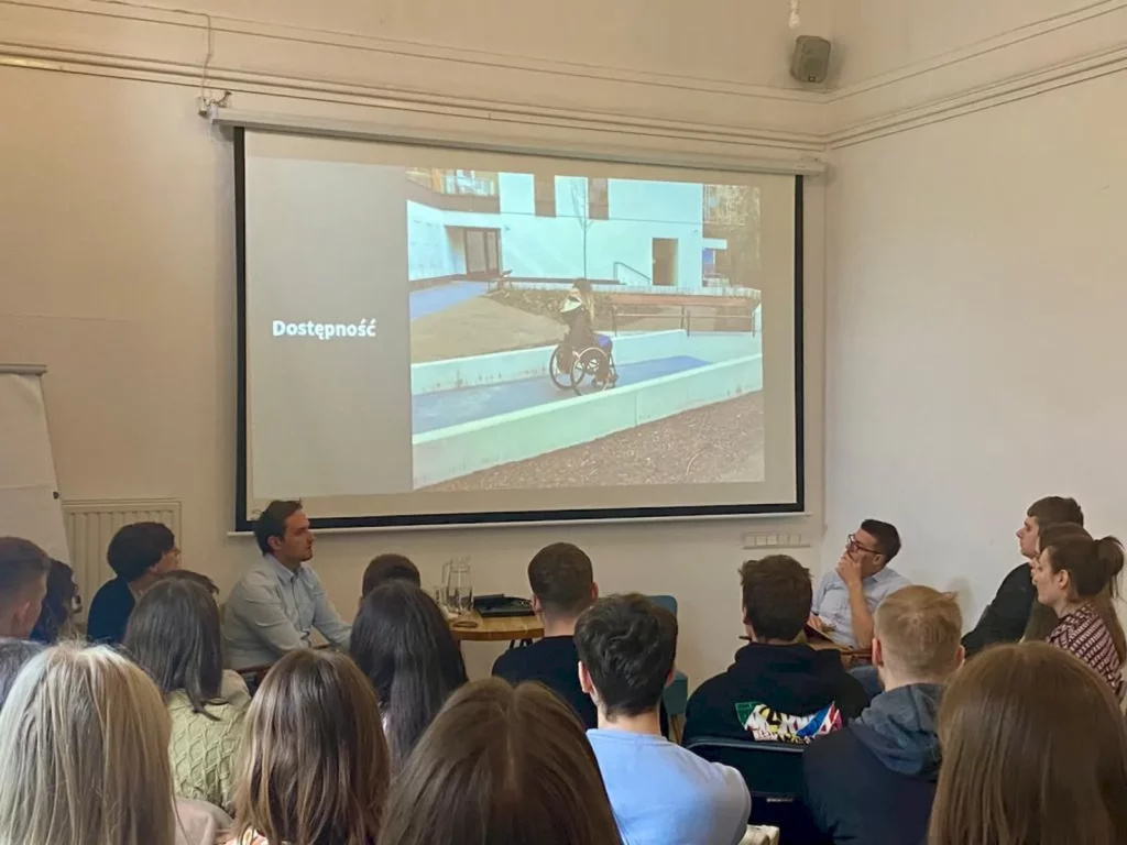 Grupa ludzi siedząca w sali konferencyjnej lub klasie, zwrócona w stronę ekranu projekcyjnego. Na ekranie wyświetlane jest zdjęcie przedstawiające osobę na wózku inwalidzkim, poruszającą się po rampie przed budynkiem. Obok zdjęcia na ekranie widnieje tekst "Dostępność". W pomieszczeniu znajduje się kilkanaście osób, które wydają się być zaangażowane w prezentację lub dyskusję. W tle widać białą tablicę.