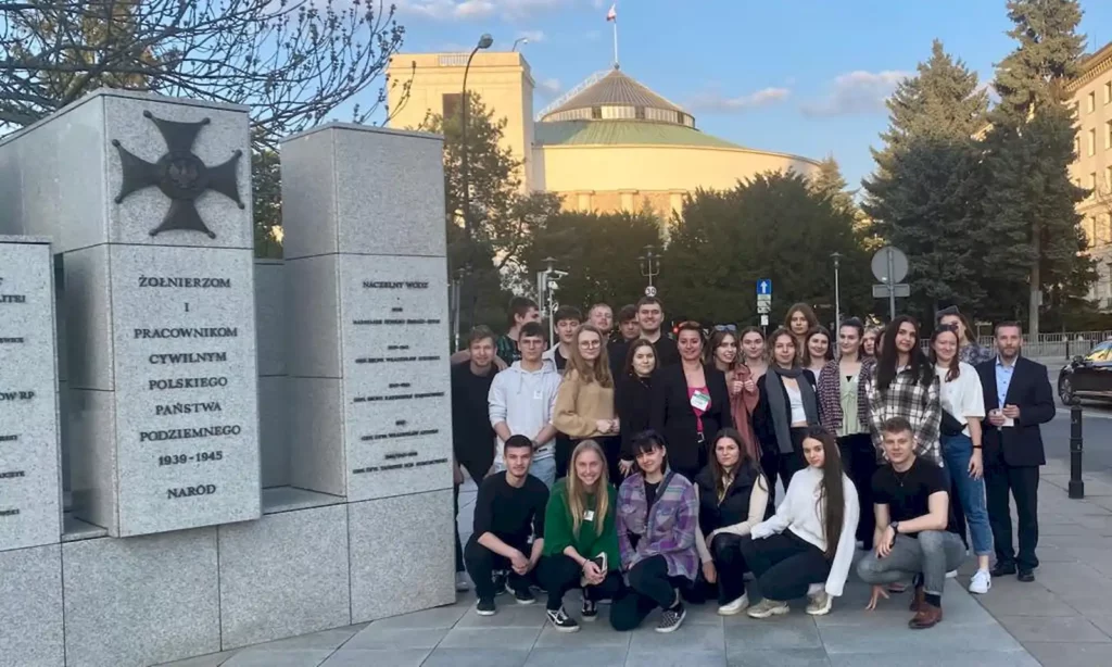 Grupa młodych ludzi stoi przed pomnikiem z kamienia, na którym widnieją napisy. W tle widać budynek z kopułą, który jest otoczony drzewami. Na szczycie budynku powiewa flaga. Pogoda jest słoneczna, a niebo jest częściowo zachmurzone. Grupa składa się z około trzydziestu osób, które pozują do zdjęcia, niektóre z nich kucają z przodu, a reszta stoi z tyłu.