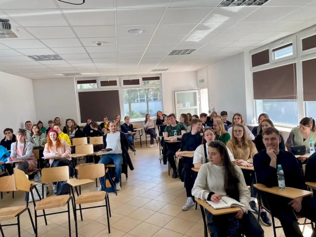 Sala konferencyjna z grupą ludzi siedzących na krzesłach, zwróconych w stronę dwóch osób stojących z przodu. Osoby te prowadzą prezentację lub wykład. W pomieszczeniu znajduje się projektor i ekran, a na ścianie wiszą portrety. Uczestnicy są różnej płci i wieku, a sala jest dobrze oświetlona. Podłoga ma kolorowe pasy, a okna są zasłonięte żaluzjami.