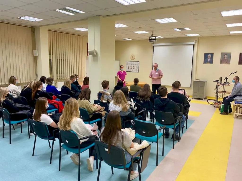 Sala konferencyjna z grupą ludzi siedzących na krzesłach, skierowanych w stronę dwóch osób stojących z przodu. Osoby te prowadzą prezentację lub wykład. W tle widać ekran projekcyjny, a na ścianie wiszą portrety. Pomieszczenie jest dobrze oświetlone, z jasnymi ścianami i podłogą w kolorze niebieskim i żółtym. Po prawej stronie znajduje się stojak z mikrofonem i sprzętem audio.