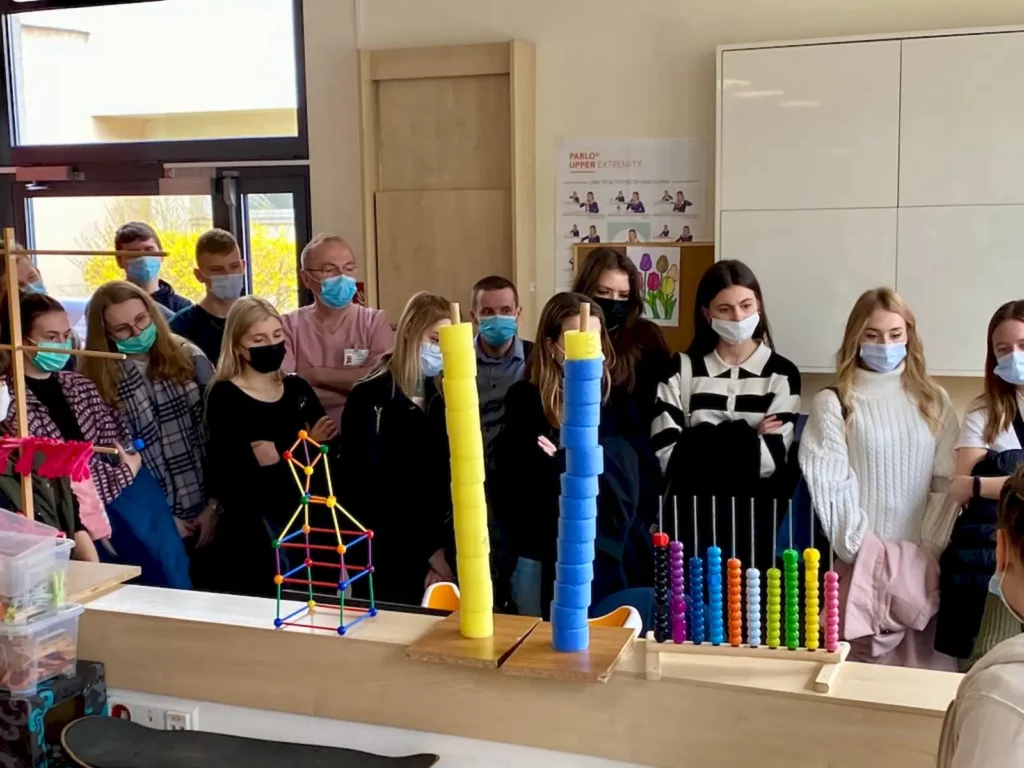 Grupa ludzi stoi w pomieszczeniu, prawdopodobnie w klasie lub sali edukacyjnej. Wszyscy mają na sobie maseczki ochronne. Przed nimi na stole znajdują się różne kolorowe pomoce dydaktyczne, takie jak wieże zbudowane z kolorowych kubków oraz zestaw kolorowych patyczków umieszczonych pionowo. W tle widać okno i drzwi, a na ścianie wiszą plakaty z ilustracjami. Grupa składa się głównie z młodych osób, prawdopodobnie uczniów, oraz kilku dorosłych, którzy mogą być nauczycielami lub opiekunami.