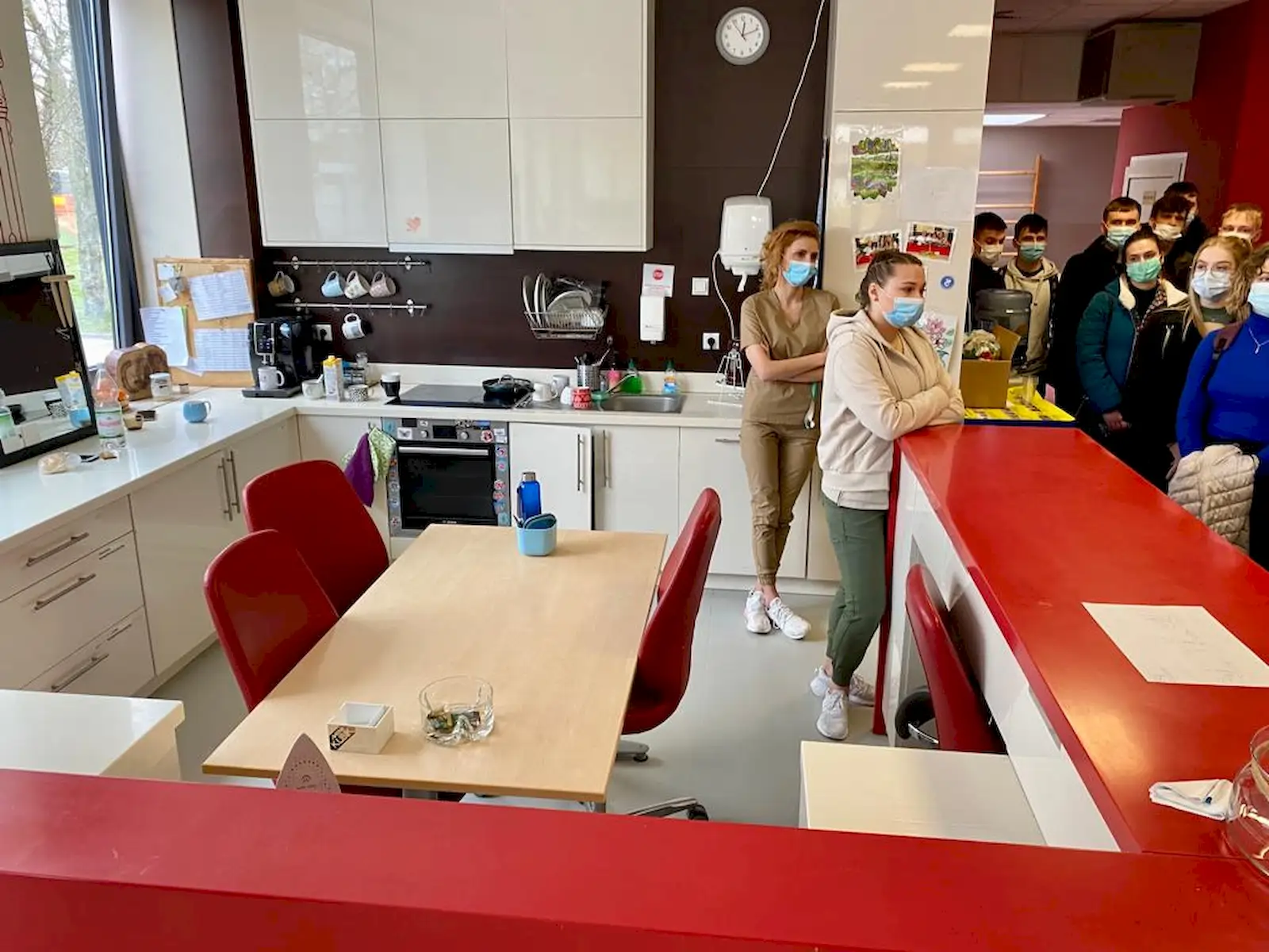 Kuchnia z nowoczesnym wystrojem. Na pierwszym planie znajduje się stół z dwoma czerwonymi krzesłami. W tle widać białe szafki kuchenne, piekarnik i zlew. Na blacie stoją różne przedmioty, w tym ekspres do kawy. Na ścianie wisi zegar. Po prawej stronie, za czerwonym blatem, stoi grupa ludzi, wszyscy noszą maseczki ochronne. Dwie osoby opierają się o blat, a reszta stoi w tle. Przez okno po lewej stronie wpada naturalne światło.