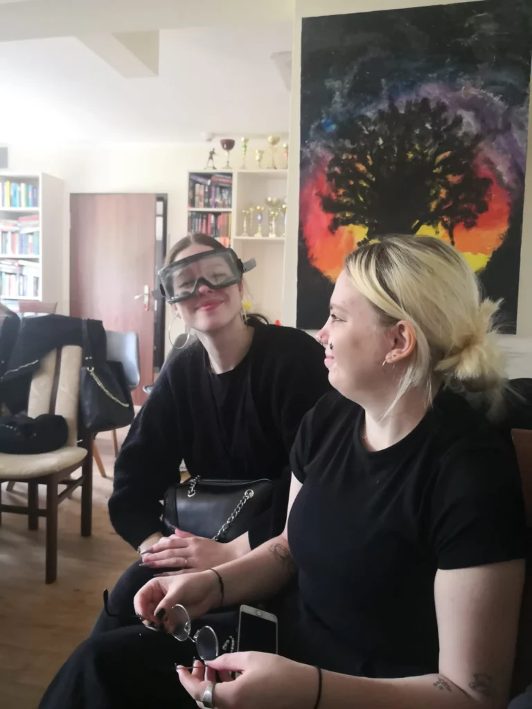 Dwie młode kobiety siedzą w pokoju. Kobieta po lewej stronie ma na sobie czarne okulary ochronne i uśmiecha się, patrząc na drugą kobietę. Kobieta po prawej stronie ma jasne włosy związane w kucyk i również się uśmiecha. Obie są ubrane na czarno. W tle widać kolorowy obraz przedstawiający drzewo na tle zachodzącego słońca oraz półki z książkami i trofeami. Pokój jest jasny i przestronny.