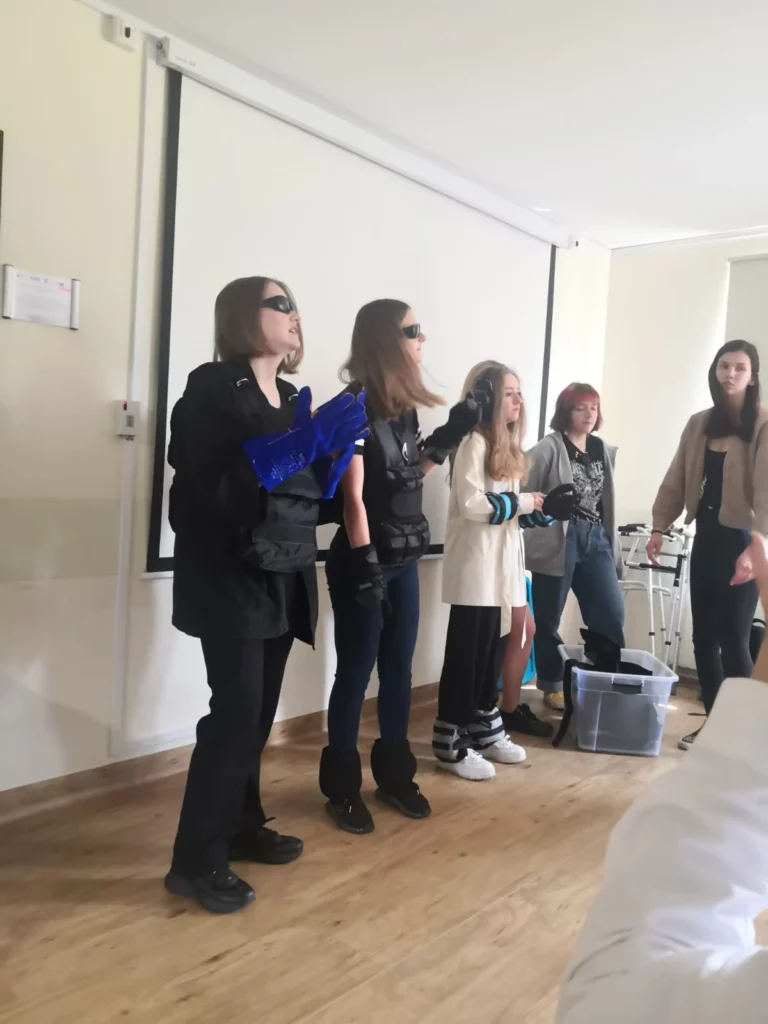 Grupa młodych osób stoi przed białym ekranem projekcyjnym w jasnym pomieszczeniu. Wszyscy mają na sobie różne stroje, a dwie osoby po lewej stronie noszą ciemne okulary  Jedna z tych osób trzyma niebieskie kartki. Osoba w środku ma na sobie białą bluzę i ciemne spodnie, a obok niej stoi osoba, ubrana w ciemne ubranie. Po prawej stronie znajduje się jeszcze jedna osoba, ubrana w jasny płaszcz. W tle widać fragmenty mebli i sprzętu, co sugeruje, że może to być sala lekcyjna lub konferencyjna.