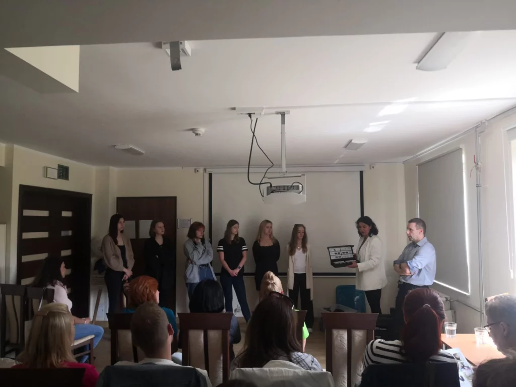 Grupa ludzi w sali konferencyjnej. Na pierwszym planie widoczni są uczestnicy siedzący na krzesłach, zwróceni w stronę prezentacji. Na środku pokoju stoi siedem osób Na suficie zamontowany jest projektor, a na ścianie za grupą znajduje się ekran projekcyjny. Pomieszczenie jest jasne, z dużymi oknami po prawej stronie.
