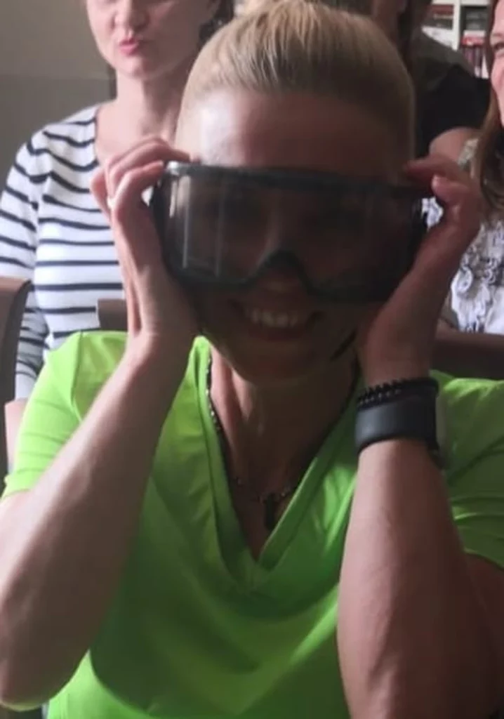 Kobieta w jasnej zielonej koszulce siedzi w pomieszczeniu i ma na sobie duże, ciemne gogle. Uśmiecha się szeroko i trzyma gogle obiema rękami. W tle widać inne osoby, z których jedna ma na sobie koszulkę w czarno-białe paski. Pomieszczenie jest dobrze oświetlone naturalnym światłem.