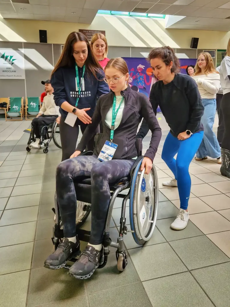 Grupa osób w pomieszczeniu. W centrum znajduje się młoda kobieta na wózku inwalidzkim, ubrana w ciemną kurtkę i spodnie. Ma na sobie identyfikator zawieszony na smyczy. Dwie kobiety stoją obok niej, pomagając jej w poruszaniu się na wózku. Jedna z nich ma na sobie czarną bluzę i niebieskie legginsy, a druga białą bluzkę. W tle widać inne osoby, w tym mężczyznę na wózku inwalidzkim oraz kilka osób stojących. Na ścianach wiszą plakaty i banery. Pomieszczenie jest dobrze oświetlone, z dużymi oknami na suficie.