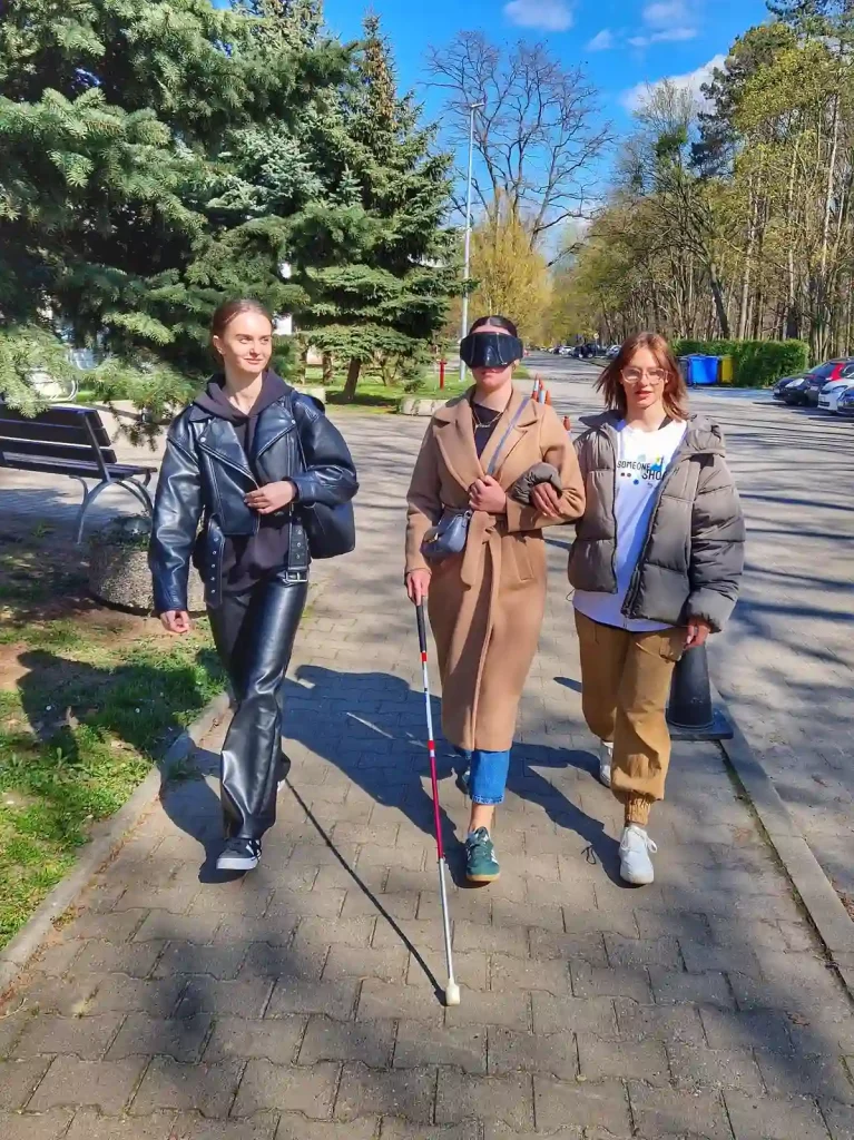 Trzy kobiety spacerujące chodnikiem w parku. Kobieta pośrodku nosi długi beżowy płaszcz, niebieskie dżinsy i czapkę z daszkiem. Trzyma białą laskę, co sugeruje, że jest osobą niewidomą. Kobieta po lewej stronie ma na sobie czarną skórzaną kurtkę i spodnie, a kobieta po prawej nosi szarą kurtkę i beżowe spodnie. W tle widać drzewa, ławkę i niebieskie niebo z kilkoma chmurami.