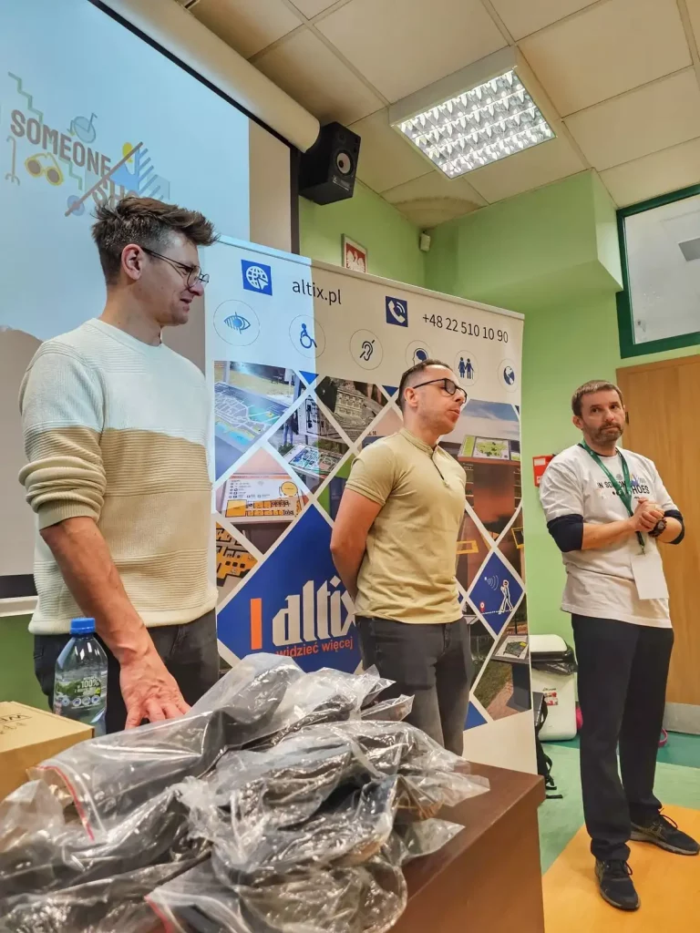 Trzech mężczyzn stoi w pomieszczeniu, prawdopodobnie podczas prezentacji lub spotkania. Dwóch z nich stoi przed roll-upem z logo "altix.pl" oraz różnymi symbolami, takimi jak ikony osób niepełnosprawnych. Na roll-upie widnieje także numer telefonu +48 22 510 10 90. Mężczyzna po lewej stronie ma na sobie jasny sweter i okulary, środkowy mężczyzna nosi jasną koszulkę i również ma okulary, a mężczyzna po prawej stronie ma na sobie białą koszulkę z zielonym nadrukiem i identyfikator na szyi. Na stole przed nimi znajdują się zapakowane w folię przedmioty oraz butelka wody. W tle widać ekran z logo "SOMEONE". Pomieszczenie ma jasnozielone ściany i sufit z białymi panelami.