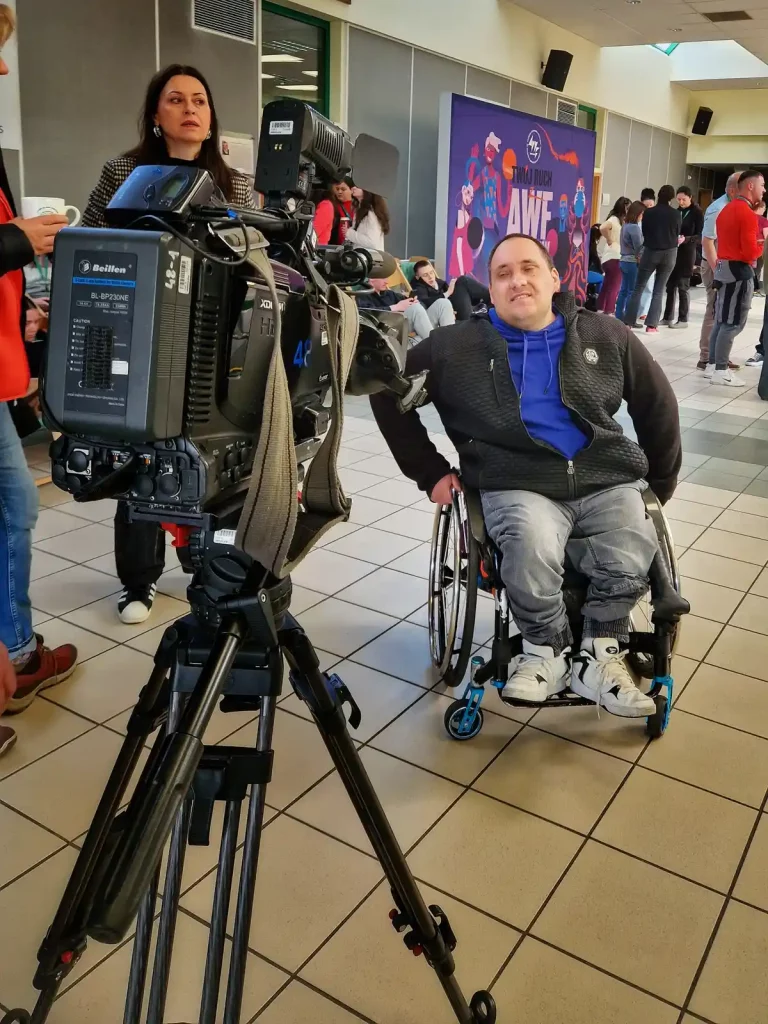 Mężczyzna na wózku inwalidzkim siedzi przed dużą kamerą telewizyjną w pomieszczeniu z kafelkową podłogą. Ma na sobie niebieską bluzę i czarną kamizelkę. W tle widać grupę ludzi, a na ścianie znajduje się kolorowy plakat z napisem "AWF". Obok kamery stoi kobieta z długimi ciemnymi włosami. Atmosfera wydaje się być związana z jakimś wydarzeniem lub konferencją.