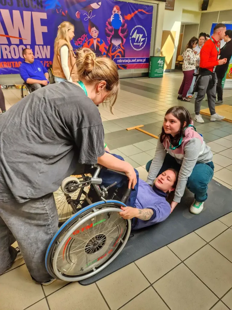 Grupa osób w pomieszczeniu, które wygląda na halę sportową lub centrum rekreacyjne. Na pierwszym planie dwie kobiety pomagają osobie na wózku inwalidzkim, która leży na macie. Jedna z kobiet trzyma wózek, a druga podtrzymuje leżącą osobę. W tle widać kilka innych osób, a na ścianie znajduje się duży plakat z napisem "AWF Wrocław" oraz grafiką przedstawiającą postacie w ruchu. Plakat promuje rekrutację na Akademię Wychowania Fizycznego we Wrocławiu.