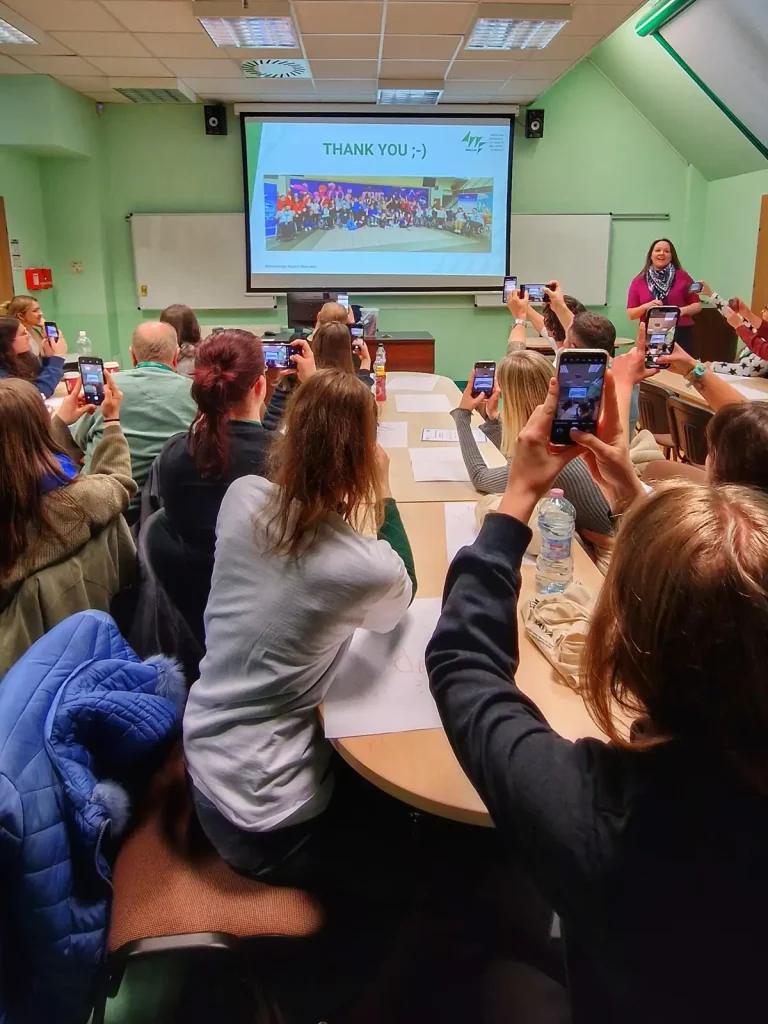 Sala wykładowa z grupą ludzi siedzących przy dużym stole. Wszyscy uczestnicy trzymają w rękach telefony komórkowe, skierowane w stronę projektora, na którym wyświetlany jest slajd z napisem "THANK YOU ;-)" oraz zdjęciem grupowym. Na pierwszym planie widać osoby siedzące tyłem do obiektywu, a na końcu sali stoi kobieta, która prowadzi prezentację. Ściany są pomalowane na jasnozielony kolor, a na stole leżą różne przedmioty, takie jak butelki z wodą i notatki.