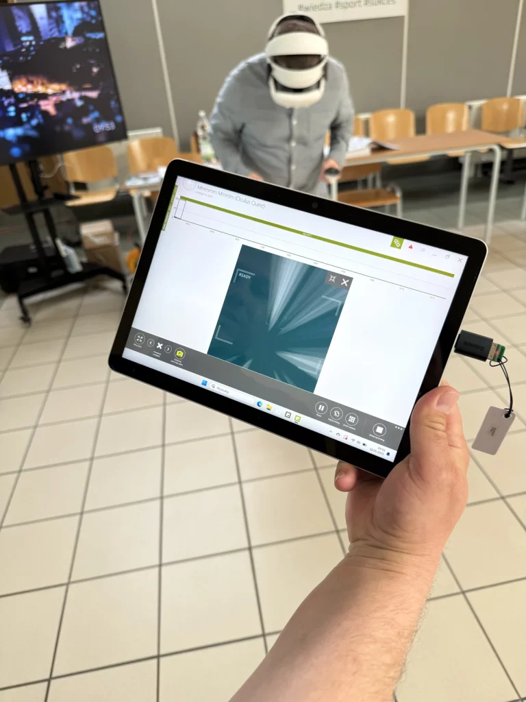 Zdjęcie przedstawia osobę noszącą zestaw do wirtualnej rzeczywistości (VR) Oculus Quest, która stoi w tle w pomieszczeniu z krzesłami i stołem. Na pierwszym planie widoczny jest tablet trzymany w ręce, na którym wyświetlany jest interfejs aplikacji związanej z VR. Na ekranie tabletu widać grafikę z napisem "READY" oraz różne elementy interfejsu użytkownika. W tle, po lewej stronie, znajduje się duży ekran, na którym wyświetlany jest rozmyty obraz. Na ścianie wisi plakat z hasztagami "#wiedza #sport #sukces". Podłoga jest wyłożona jasnymi płytkami.