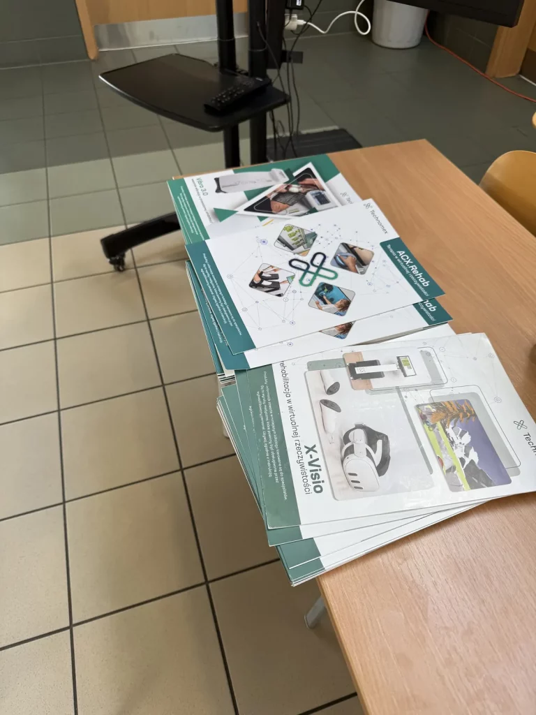 Stos ulotek leżących na stole. Ulotki mają zielone i białe elementy graficzne oraz zdjęcia przedstawiające różne urządzenia technologiczne. Na jednej z ulotek widnieje napis "XVisio" oraz tekst dotyczący rehabilitacji w wirtualnej rzeczywistości. W tle widać podłogę z płytek, część krzesła oraz stojak z monitorem i klawiaturą.