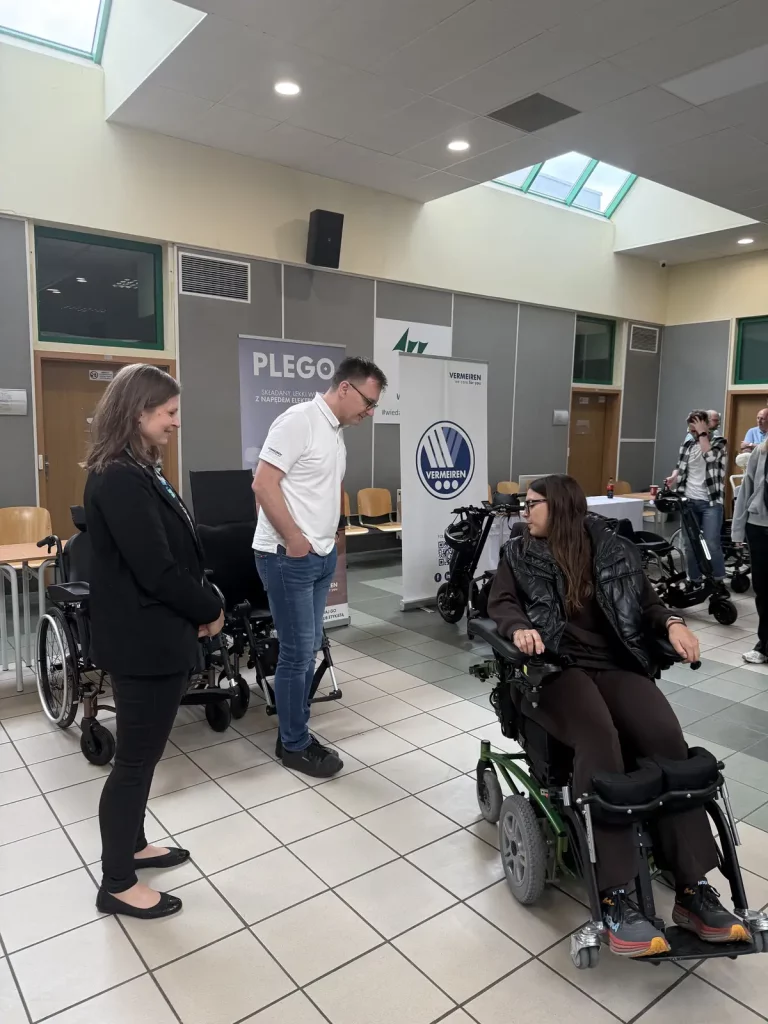 Zdjęcie przedstawia wnętrze sali, w której odbywa się prezentacja lub spotkanie dotyczące wózków inwalidzkich. Na pierwszym planie znajduje się kobieta siedząca na elektrycznym wózku inwalidzkim. Obok niej stoi mężczyzna w białej koszulce i dżinsach oraz kobieta w ciemnym stroju. W tle widać kilka plakatów i banerów, na jednym z nich widnieje napis "PLEGO". W pomieszczeniu znajduje się kilka innych osób, a także różne modele wózków inwalidzkich. Sala jest dobrze oświetlona, z dużymi oknami w suficie.