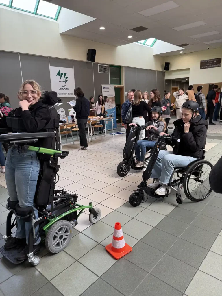 Grupa ludzi w przestronnym pomieszczeniu. Na pierwszym planie znajduje się kobieta stojąca na wózku inwalidzkim z funkcją pionizacji, uśmiechająca się do kamery. Obok niej, po prawej stronie, są dwie osoby na wózkach inwalidzkich, które również wyglądają na zadowolone. Na podłodze przed nimi stoi pomarańczowy pachołek drogowy. W tle widać stoisko z plakatem "AWF Wrocław" oraz kilka osób rozmawiających ze sobą. Pomieszczenie jest dobrze oświetlone dzięki dużym oknom dachowym.