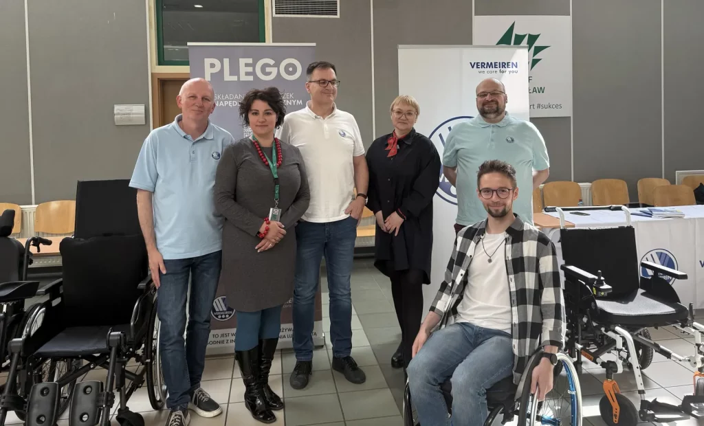 Grupa sześciu osób stoi w pomieszczeniu, a jedna osoba siedzi na wózku inwalidzkim. W tle znajdują się dwa roll-upy z napisami "PLEGO" i "VERMEIREN we care for you". Osoby są uśmiechnięte i wyglądają na zadowolone. Na pierwszym planie po lewej stronie widać dwa wózki inwalidzkie. Pomieszczenie ma szare ściany i drewniane krzesła ustawione w rzędzie. Na stole po prawej stronie leżą broszury lub dokumenty.