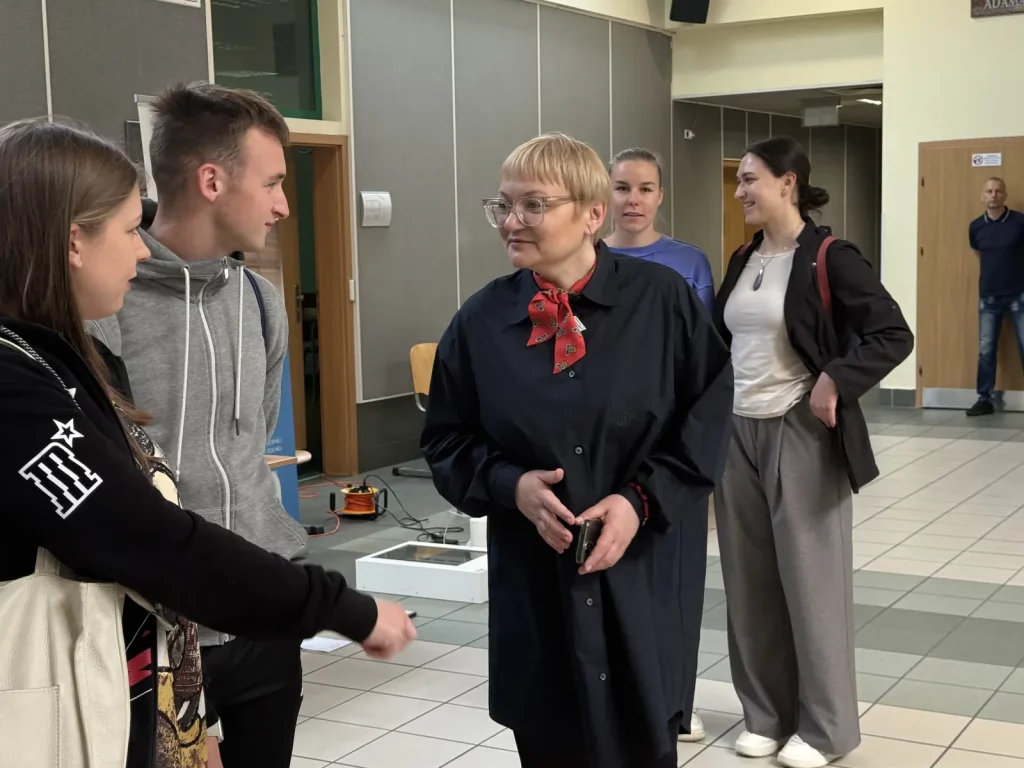 Grupa ludzi w przestronnym pomieszczeniu. Na pierwszym planie znajduje się kobieta w okularach, ubrana w ciemny płaszcz i czerwoną apaszkę w kwiaty. Rozmawia z młodym mężczyzną w szarej bluzie z kapturem oraz młodą kobietą w czarnej bluzie z białymi nadrukami. W tle widać dwie inne kobiety, jedna w fioletowej bluzce, a druga w białej koszulce i czarnej marynarce, obie uśmiechnięte. W rogu pomieszczenia stoi mężczyzna w ciemnej koszulce i jeansach. Na podłodze leżą kable i sprzęt elektroniczny. Pomieszczenie jest jasne, z szarymi ścianami i drzwiami w tle.