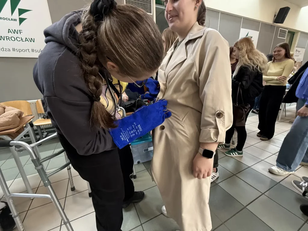 Kobieta w beżowym płaszczu stoi w pomieszczeniu, podczas gdy inna osoba, ubrana w ciemną bluzę z kapturem, pochyla się nad nią, trzymając niebieskie rękawice ochronne. W tle widać kilka innych osób, które stoją w kolejce. Na ścianie po lewej stronie znajduje się znak z napisem "AWF WROCŁAW". Pomieszczenie wygląda na przestrzeń publiczną, być może w budynku uczelni lub centrum konferencyjnym. Na podłodze są płytki, a po lewej stronie widać kilka krzeseł.