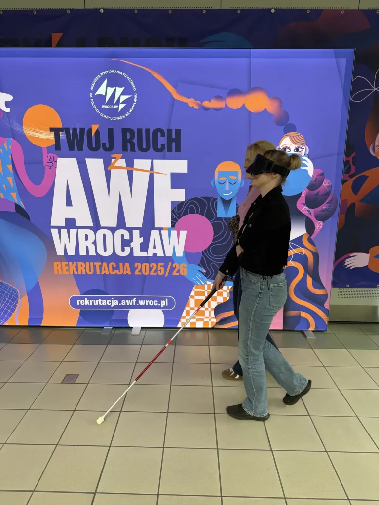 Plakat promocyjny AWF Wrocław. Na pierwszym planie znajduje się młoda kobieta z opaską na oczach, trzymająca białą laskę, prowadzona przez inną osobę. W tle widoczny jest kolorowy plakat z napisem "TWÓJ RUCH AWF WROCŁAW REKRUTACJA 2025/26". Plakat ma intensywne kolory, takie jak fiolet, pomarańcz i niebieski, oraz zawiera grafikę przedstawiającą postacie i różne kształty. Na plakacie znajduje się również adres strony internetowej: rekrutacja.awf.wroc.pl.
