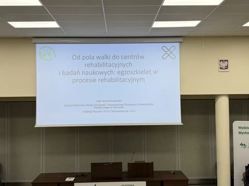 Prezentacja na ekranie w sali konferencyjnej. Na slajdzie widnieje tekst: "Od pola walki do centrów rehabilitacyjnych i badań naukowych: egzoszkielet w procesie rehabilitacyjnym". Poniżej znajduje się informacja o autorce: "mgr Anna Krzyżańska Szkoła Doktorska Klinika Ortopedii i Traumatologii Dziecięcej Uniwersytetu Medycznego w Poznaniu X-Rehab Poznań - P.H.U. Technomex sp. z o.o.". W lewym górnym rogu slajdu znajduje się logo z napisem "Poznań", a w prawym górnym rogu jest symbol przypominający skrzyżowane linie. Na ścianie obok ekranu wisi godło Polski. Na dole zdjęcia widać fragment mównicy.