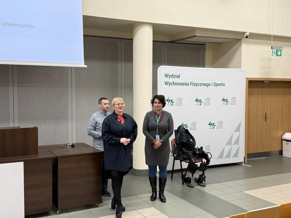 Trzy osoby stojące w sali wykładowej. Na pierwszym planie kobieta w ciemnej sukience z czerwoną apaszką, obok niej kobieta w szarej sukience z zielonym identyfikatorem na szyi. Za nimi stoi mężczyzna w szarym swetrze. W tle znajduje się biały ekran projekcyjny oraz tablica z napisem "Wydział Wychowania Fizycznego i Sportu" i logo Akademii Wychowania Fizycznego we Wrocławiu. Obok tablicy stoi krzesło z egzoszkieletem. Po prawej stronie widoczne są drzwi i kosz na śmieci.