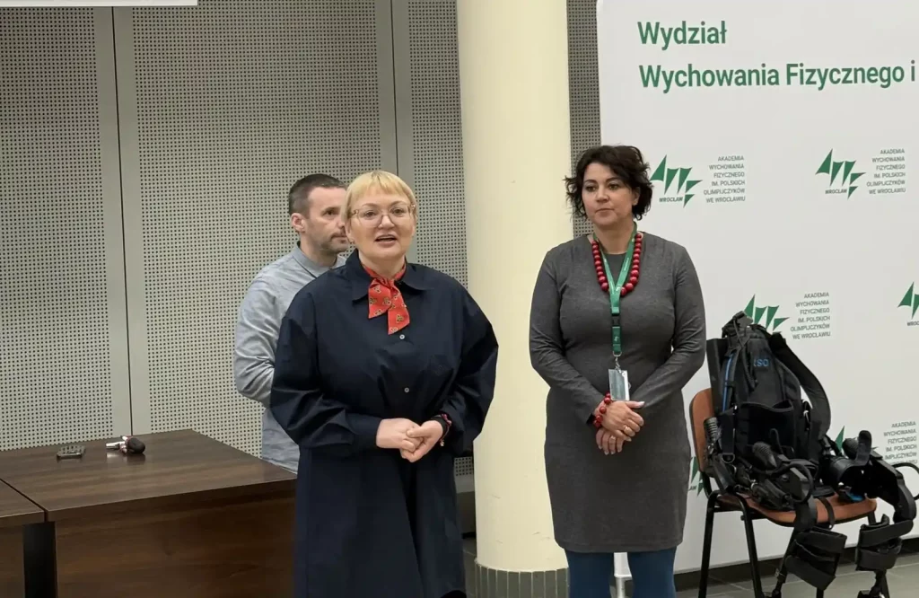 Trzy osoby stoją w pomieszczeniu, które wygląda na salę konferencyjną lub wykładową. Na pierwszym planie jest kobieta z krótkimi blond włosami, nosząca ciemny płaszcz i czerwoną apaszkę. Obok niej stoi kobieta z ciemnymi włosami, ubrana w szarą sukienkę i czerwony naszyjnik. Za nimi, nieco w tle, znajduje się mężczyzna w szarej koszuli. Na ścianie po prawej stronie widać napis "Wydział Wychowania Fizycznego i...", a poniżej logo Akademii Wychowania Fizycznego we Wrocławiu. Obok kobiet stoi krzesło, na którym znajduje się sprzęt przypominający egzoszkielet.