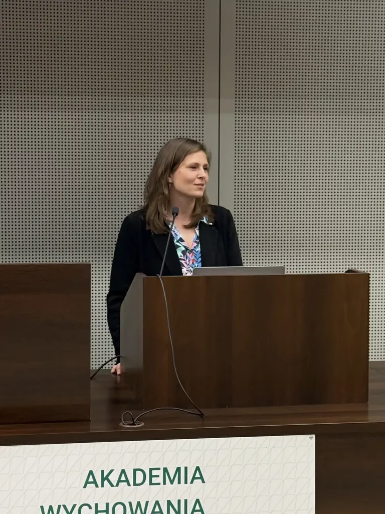 Kobieta stojąca za drewnianą mównicą w sali konferencyjnej. Ma na sobie ciemną marynarkę i kolorową bluzkę. Przed nią znajduje się mikrofon. W tle widać perforowaną ścianę. Na mównicy jest napis "AKADEMIA WYCHOWANIA".