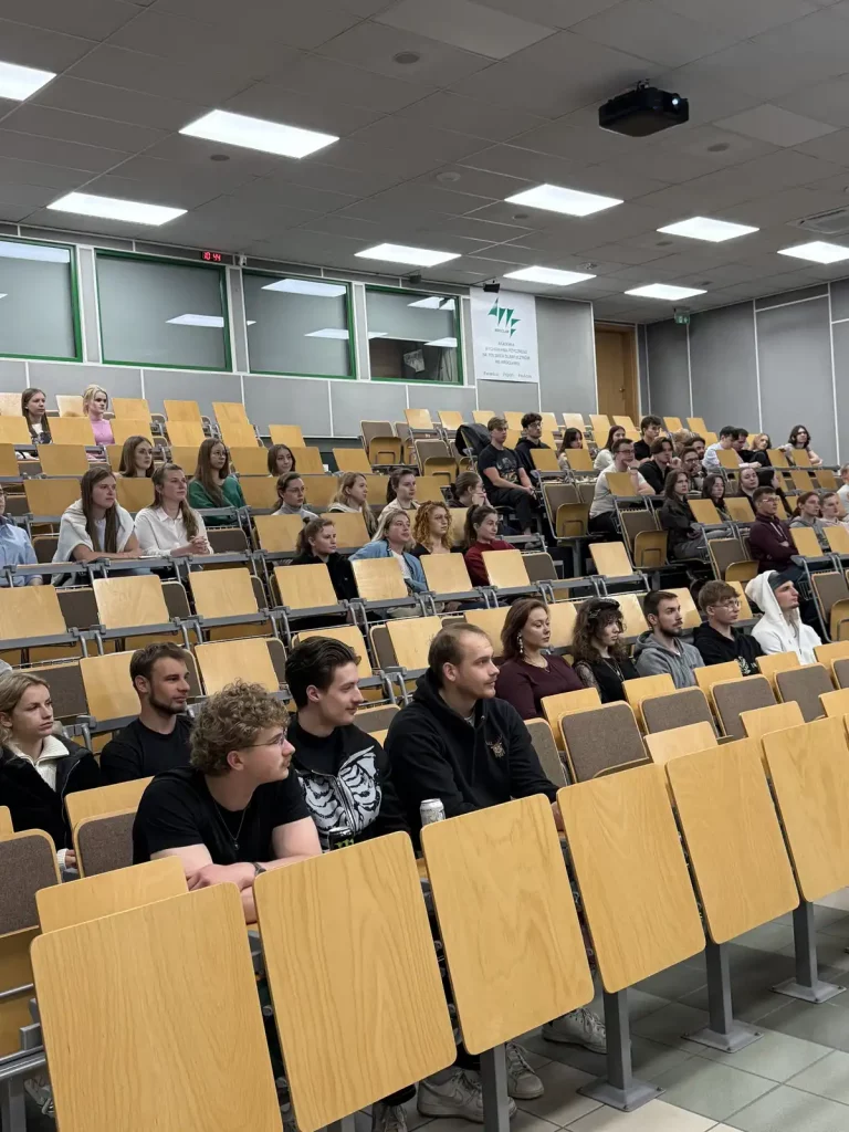 Sala wykładowa z drewnianymi siedzeniami, wypełniona studentami. Wszyscy siedzą na krzesłach ustawionych w rzędach, patrząc w stronę, gdzie prawdopodobnie znajduje się wykładowca. Na ścianie z tyłu widnieje logo i napis "Instytut Informatyki Stosowanej i Mechaniki Stosowanej". Widać również zegar pokazujący godzinę 10:54. Oświetlenie jest jasne, a sufit pokryty jest panelami z lampami. Studenci są różnorodnie ubrani, niektórzy mają na sobie ciemne ubrania, inni jaśniejsze.