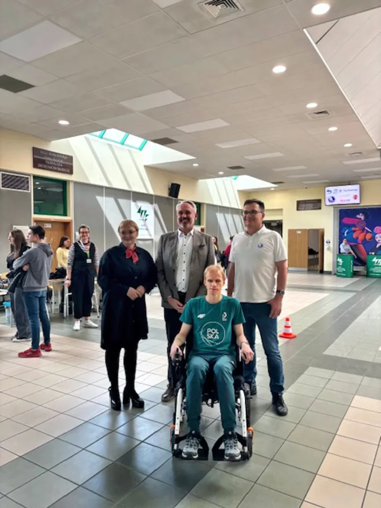 Grupa czterech osób w przestronnym, jasnym holu budynku. Na pierwszym planie siedzi młoda osoba na wózku inwalidzkim, ubrana w zielony sportowy dres z napisem "POLSKA" na koszulce. Za nią stoją trzy osoby: kobieta w ciemnym stroju z czerwoną apaszką oraz dwóch mężczyzn w jasnych koszulach i marynarkach. W tle widać kilka innych osób, jasne ściany, drzwi oraz tablice informacyjne. Pomieszczenie jest dobrze oświetlone światłem dziennym wpadającym przez duże okna.
