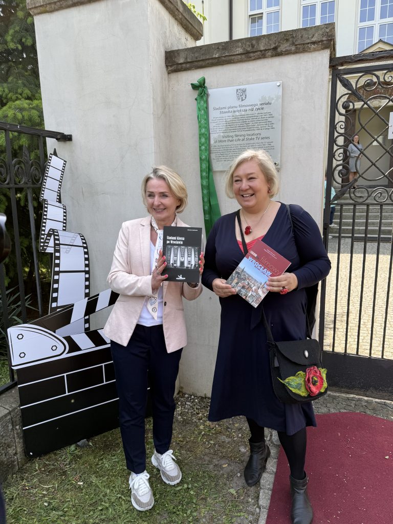 Dwie kobiety stojące na zewnątrz przed bramą. Obie trzymają książki i uśmiechają się do kamery. Kobieta po lewej stronie ma na sobie jasną marynarkę, ciemne spodnie i białe buty, a w rękach trzyma książkę z czarną okładką. Kobieta po prawej stronie ubrana jest w ciemną sukienkę i trzyma książkę z czerwoną okładką. Na ścianie za nimi znajduje się tablica pamiątkowa, a obok nich stoi dekoracja w kształcie klapsa filmowego. Na ziemi leży czerwona mata.