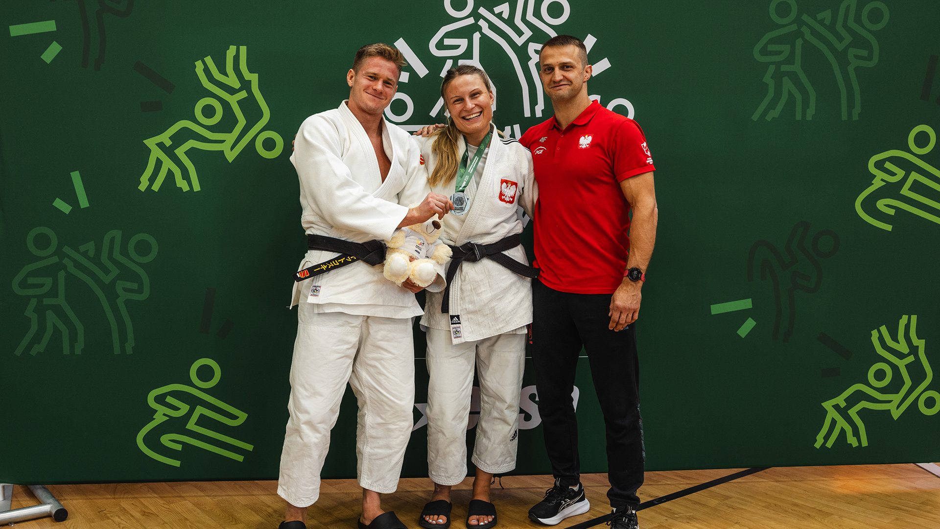 dwie zawodniczki judo w białych strojach z czarnymi pasami i trener