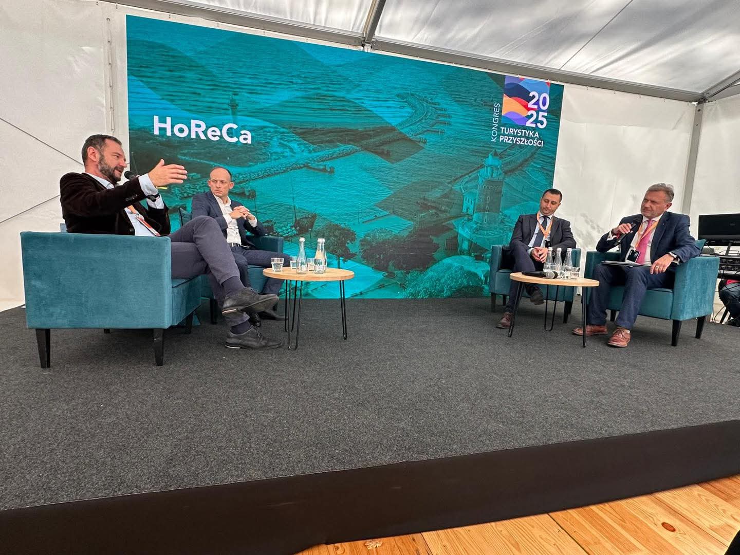 panel dyskusyjny na scenie, mężczyźni siedzą w fotelach na tle ścianki reklamowej