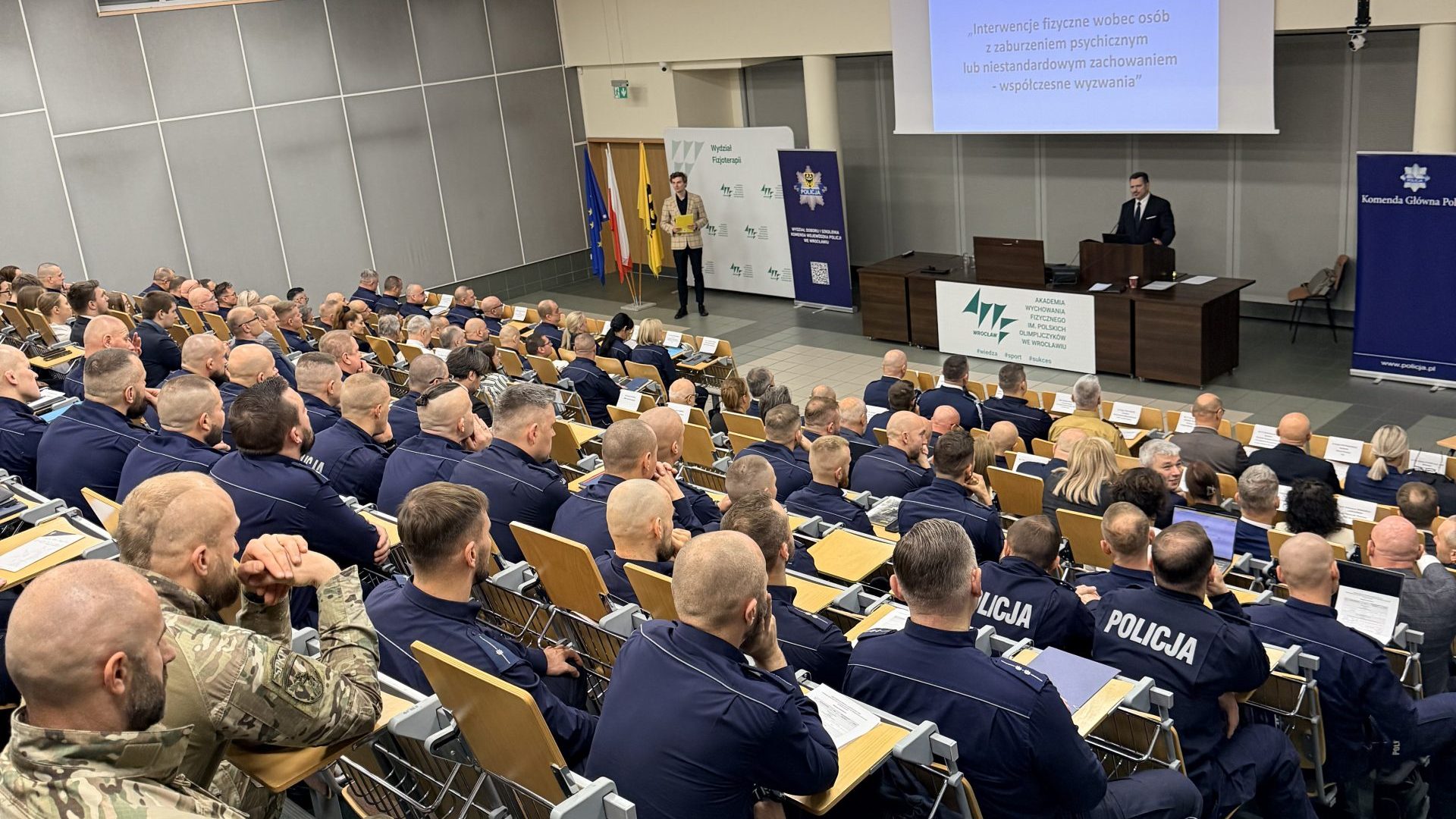 sala wykładowa zapełniona uczestnikami konferencji, na dole rektor otwierający konferencję